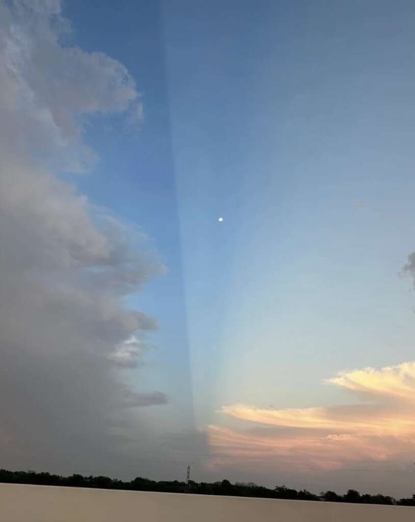 A serene sky at dusk with the moon visible. To the left, dark storm clouds contrast with a clear blue sky on the right, reflecting subtle pink and orange hues near the horizon. A faint beam of light stretches upward in the center.