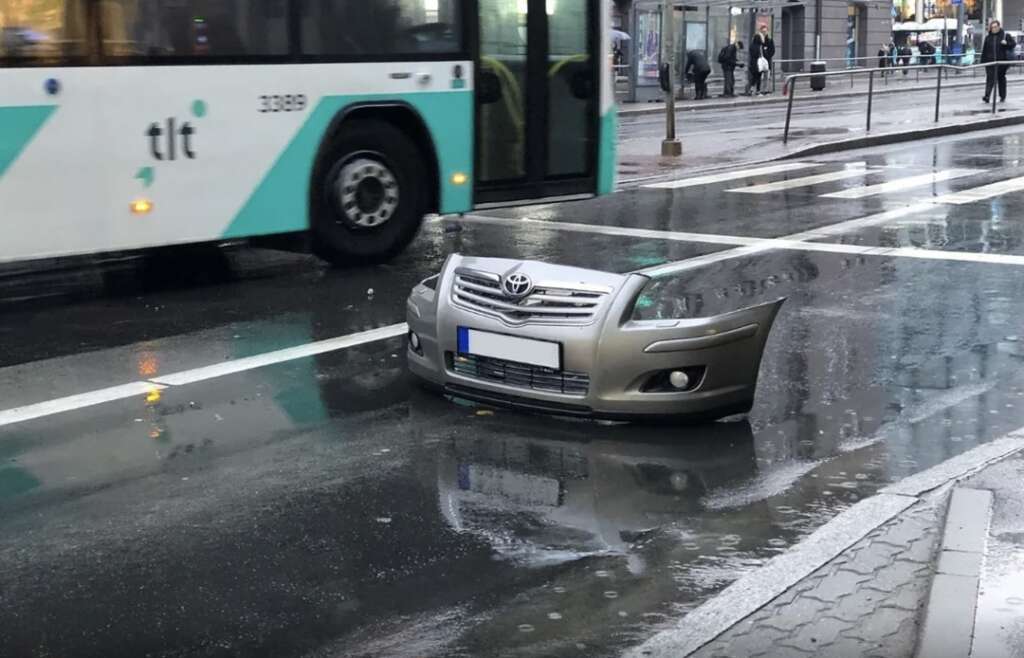 A detached car bumper lying in the middle of a wet street, close to a moving bus. The street is shiny from rain, and pedestrians can be seen in the background.