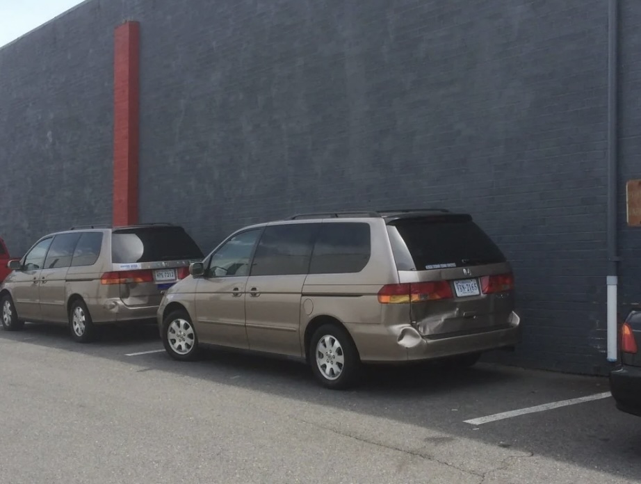 Two beige minivans are parked next to each other in a parking lot, adjacent to a tall, plain gray wall. A red stripe runs vertically along part of the wall. The scene is calm and overcast.