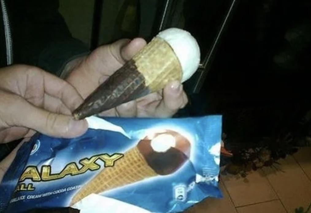 A person holding an ice cream cone with the scoop positioned incorrectly at the tip of the cone. The packaging shown reads "Galaxy" and displays a picture of a normal ice cream cone.