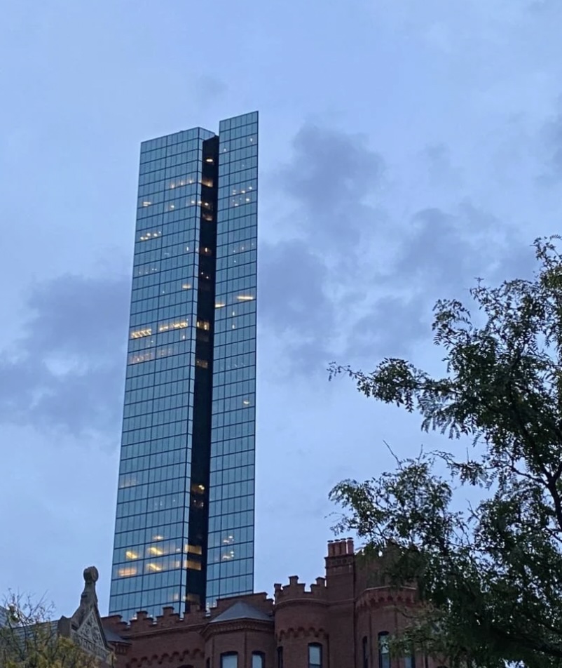 Tall glass skyscraper with illuminated windows against a cloudy evening sky, rising behind a red-brick building with ornate architectural details and a tree in the foreground.