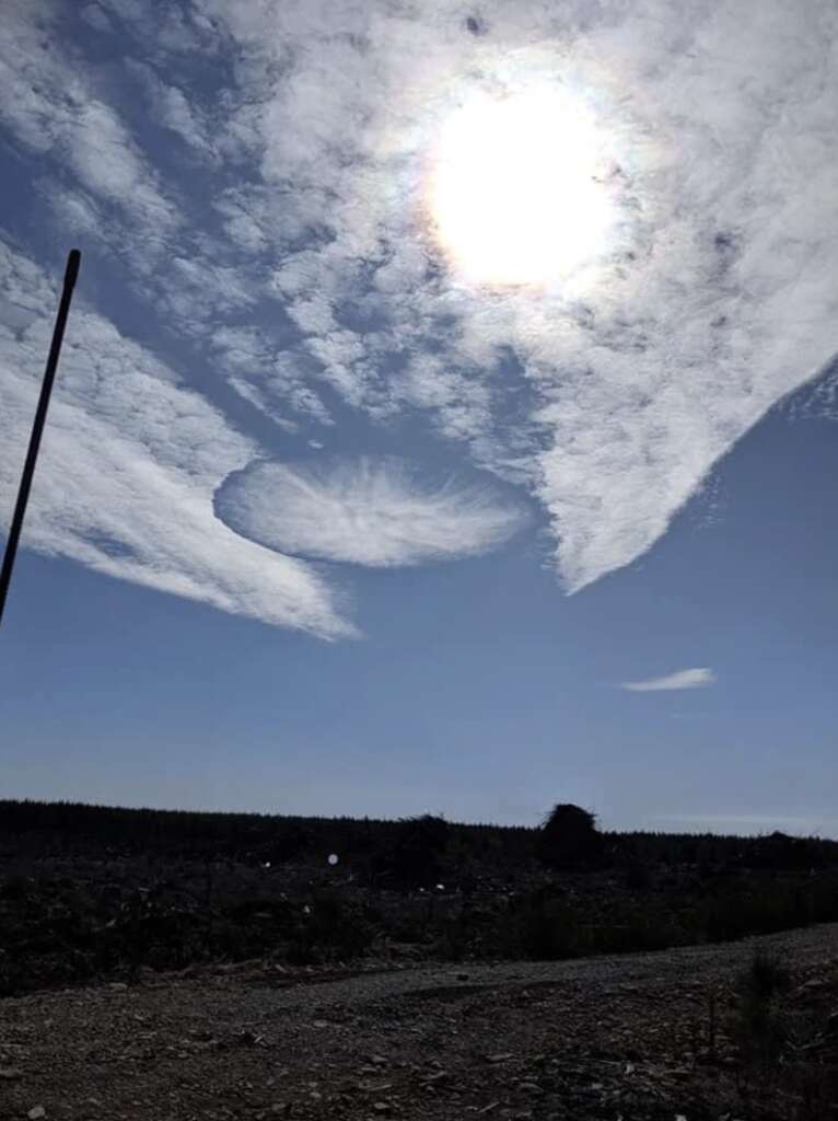 A bright sun shines in a blue sky with scattered clouds. A distinct hole-punch cloud formation is visible, creating an unusual circular gap amidst the clouds. The foreground shows a silhouette of land and a vertical pole.