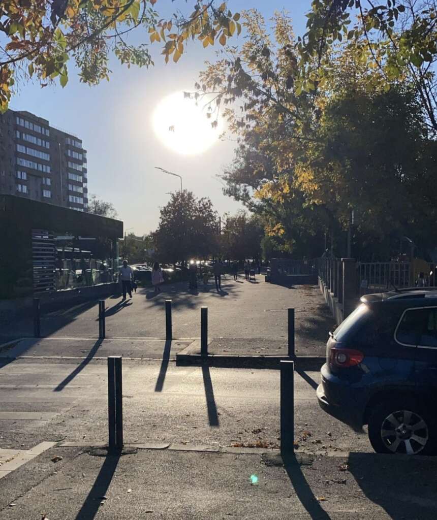 The image shows a sunny street scene in the late afternoon. The sun is low, casting long shadows. Trees with autumn leaves line the sidewalk. A blue car is parked on the right, and pedestrians are in the distance near a modern building.