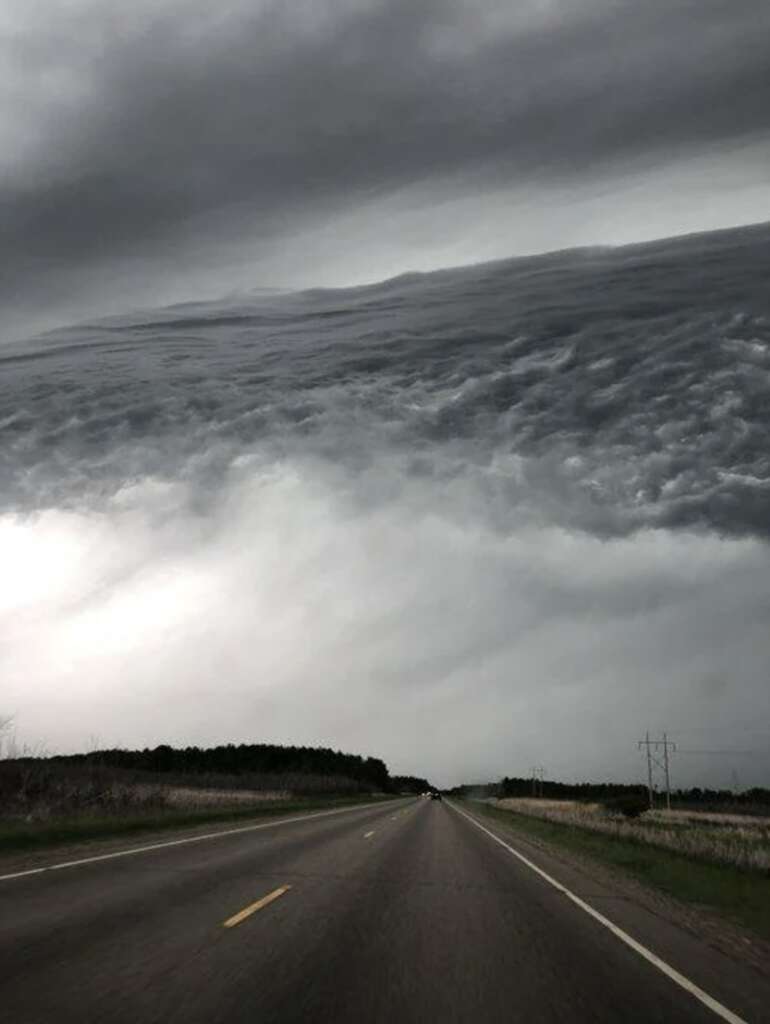 A rural road leads into the distance under a dramatic sky filled with thick, swirling gray clouds. The horizon is lined with dark trees, and the ominous sky dominates the scene, creating a sense of impending weather change.