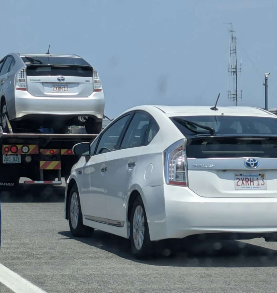 Two identical white Toyota Prius cars are on a road; one is on a flatbed truck, and the other is driving behind. Both cars have the same license plate number: 2XRH13. The background shows a clear sky and a tall communication tower.