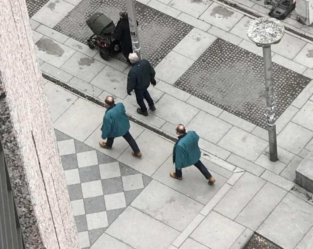 Two identical men in blue jackets and brown boots walk on a city sidewalk, passing by an older man pushing a stroller. The street is paved with geometric tiles and has modern lampposts. The scene is viewed from above.