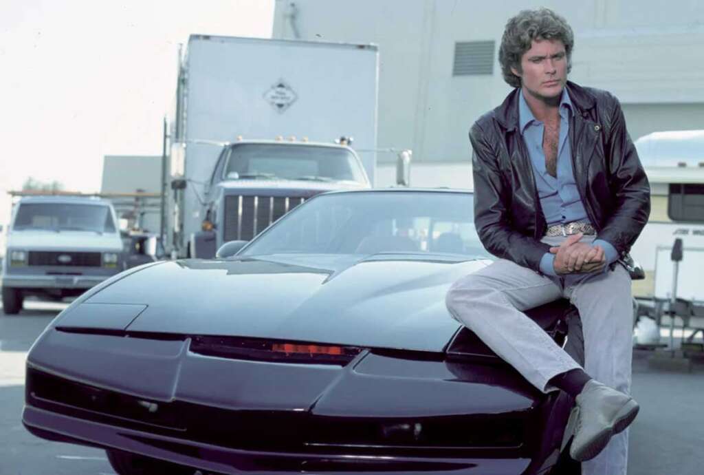 A person in a leather jacket and jeans sits on the hood of a sleek black car with a futuristic red light, reminiscent of the coolest cars from the 1980s. Trucks are parked in the background.