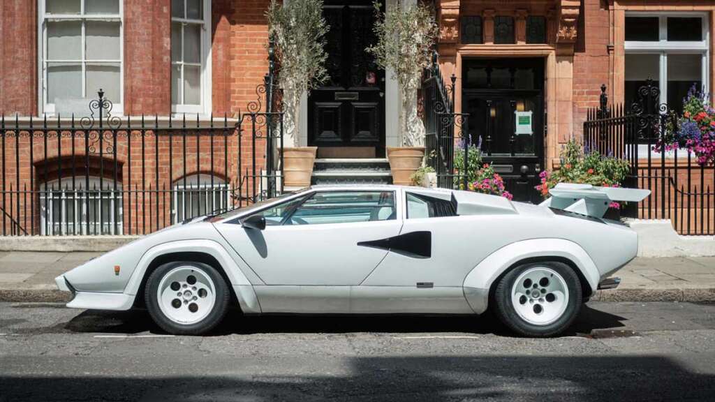 A classic white sports car, reminiscent of the coolest cars from the 1980s, is parked on a city street in front of red-brick buildings. It features a distinctive angular design and a large rear wing. Potted plants and flowers adorn the building entrances.