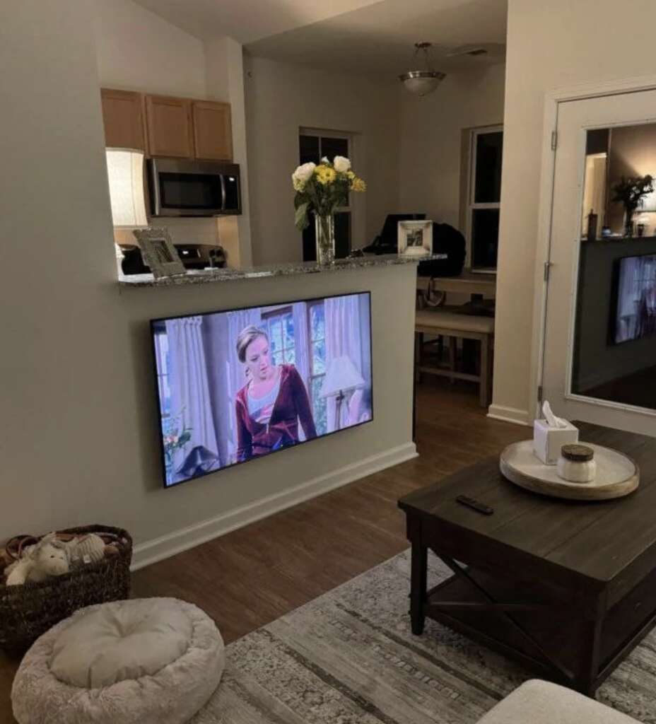 A cozy living room features a wall-mounted TV displaying a mildly infuriating scene with a person. There's a round pouf, a coffee table, and a cat bed with a sleeping cat. In the background, the kitchen bar adorned with flowers completes the setting.