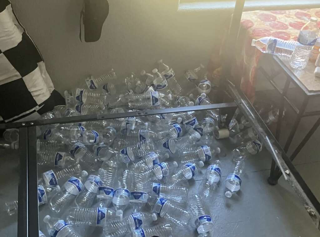 A mildly infuriating pile of empty plastic water bottles is scattered on the floor under and around a bed frame in a sunlit room. A portion of a checkered blanket and a colorful bedspread peek out from the left and right.