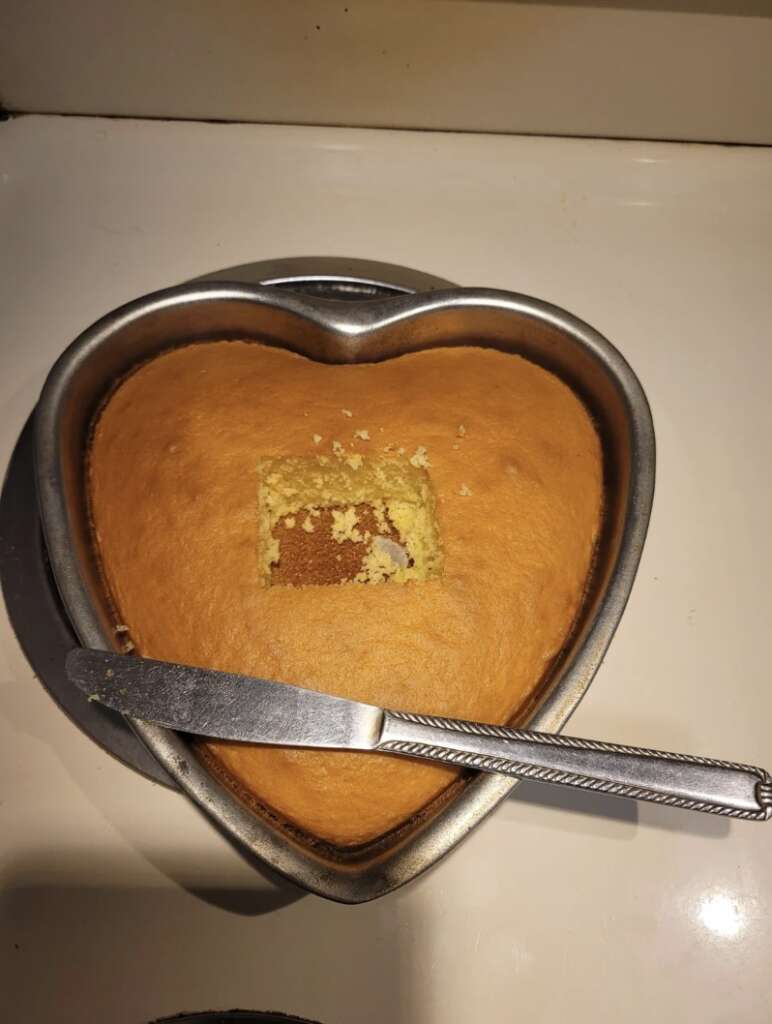 A heart-shaped cake with a square piece cut out, resting in a pan, sits mildly infuriatingly on the surface. A knife with a textured handle teeters at the pan's edge. The light brown cake contrasts starkly against its white backdrop, adding to the scene's quirky imperfection.