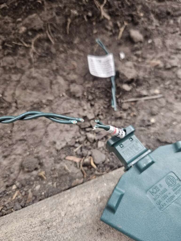 Close-up of a cut string of green outdoor holiday lights on the ground, with exposed wires and a plastic label nearby—an undeniably mildly infuriating sight. The background shows dirt and some debris.