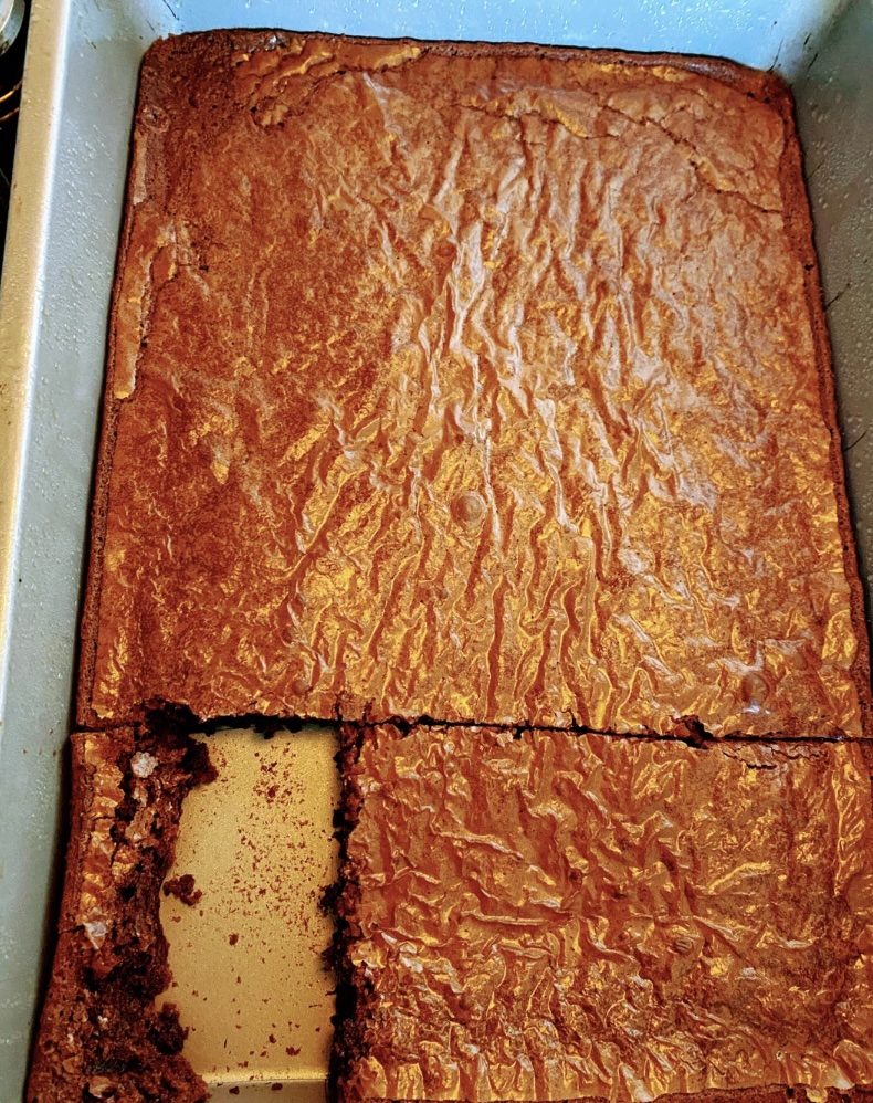 A baking pan with freshly baked brownies sits tantalizingly on the counter. One section has been cut and removed, revealing a dense and fudgy interior, yet it's mildly infuriating how the shiny, crackly top remains perfectly intact everywhere else.