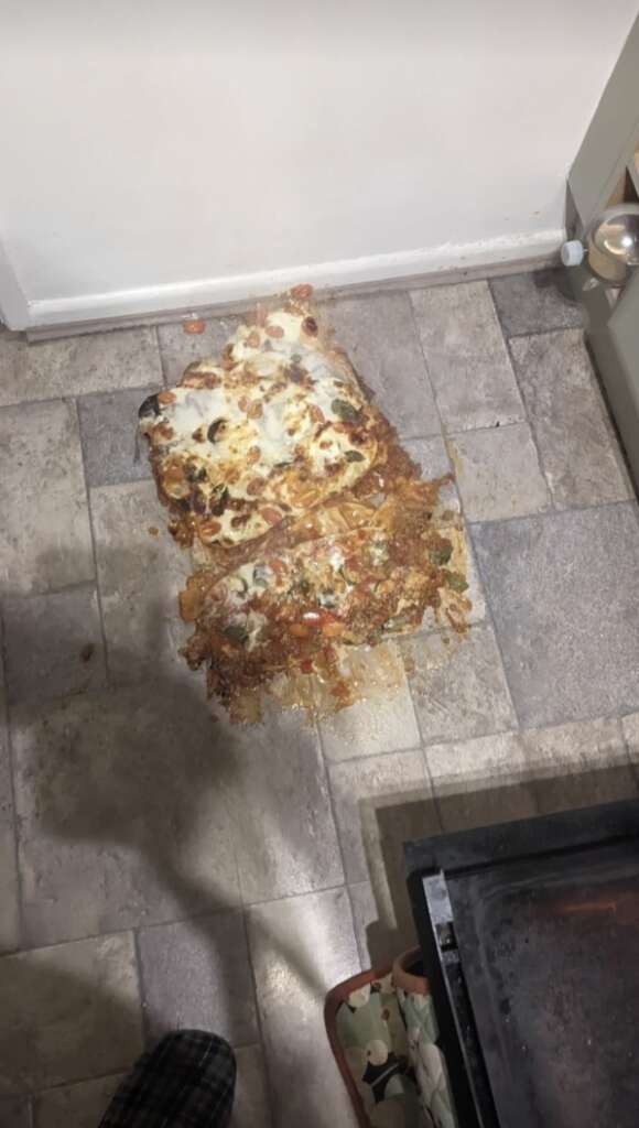 A homemade pizza has fallen face-down on a kitchen floor in an mildly infuriating scene. Toppings and melted cheese are scattered, with part of the sauce smeared on the tiles, while the oven door stands slightly open nearby.