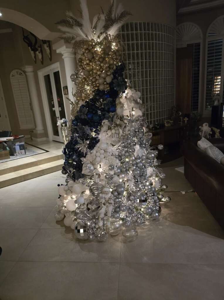 A festive Christmas tree adorned with silver, blue, and white ornaments, feathers, and lights stands in a dimly lit room. Its mildly infuriating spiral design places baubles at odd angles, creating an uneven yet sparkling effect that captivates the eye.