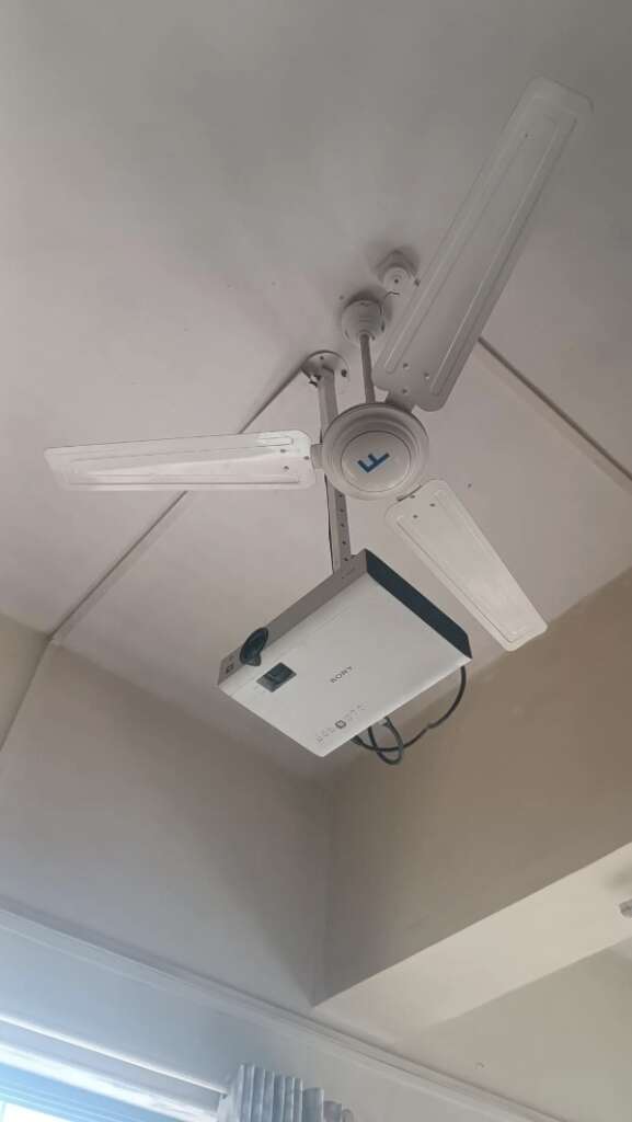 A mildly infuriating ceiling fan with three blades features a projector awkwardly mounted underneath. The setup, attached to a white ceiling, offers an incomplete view of a window and curtain, adding to the peculiar ambiance.