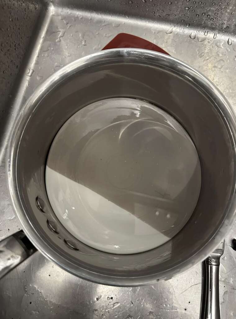 A top-down view of a metal pot with water inside, awkwardly placed in a stainless steel sink. Part of a red object is teasingly visible near the top, while the pot's handle frustratingly juts out on the left side. Reflections and shadows dance mockingly on the water's surface—mildly infuriating!