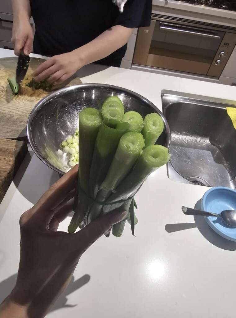 A person holds a bunch of cut celery stalks in the kitchen, while another chops ingredients near a metal bowl with chopped vegetables. Meanwhile, the mildly infuriating chaos of ingredients scattered around adds to the scene, complemented by a sink and a blue bowl in the background.