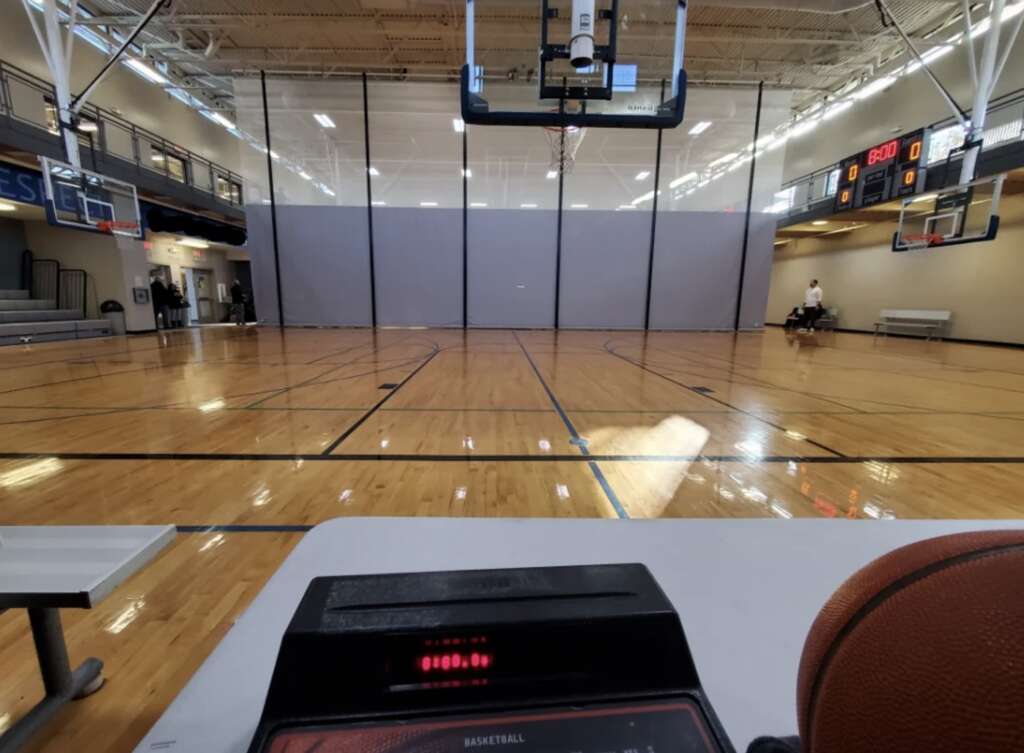 A gymnasium with polished wooden floors and multiple basketball hoops. A scoreboard, mildly infuriating in its finality, shows a score of 96 to 100. A basketball sits on a table beside a digital display near the foreground.