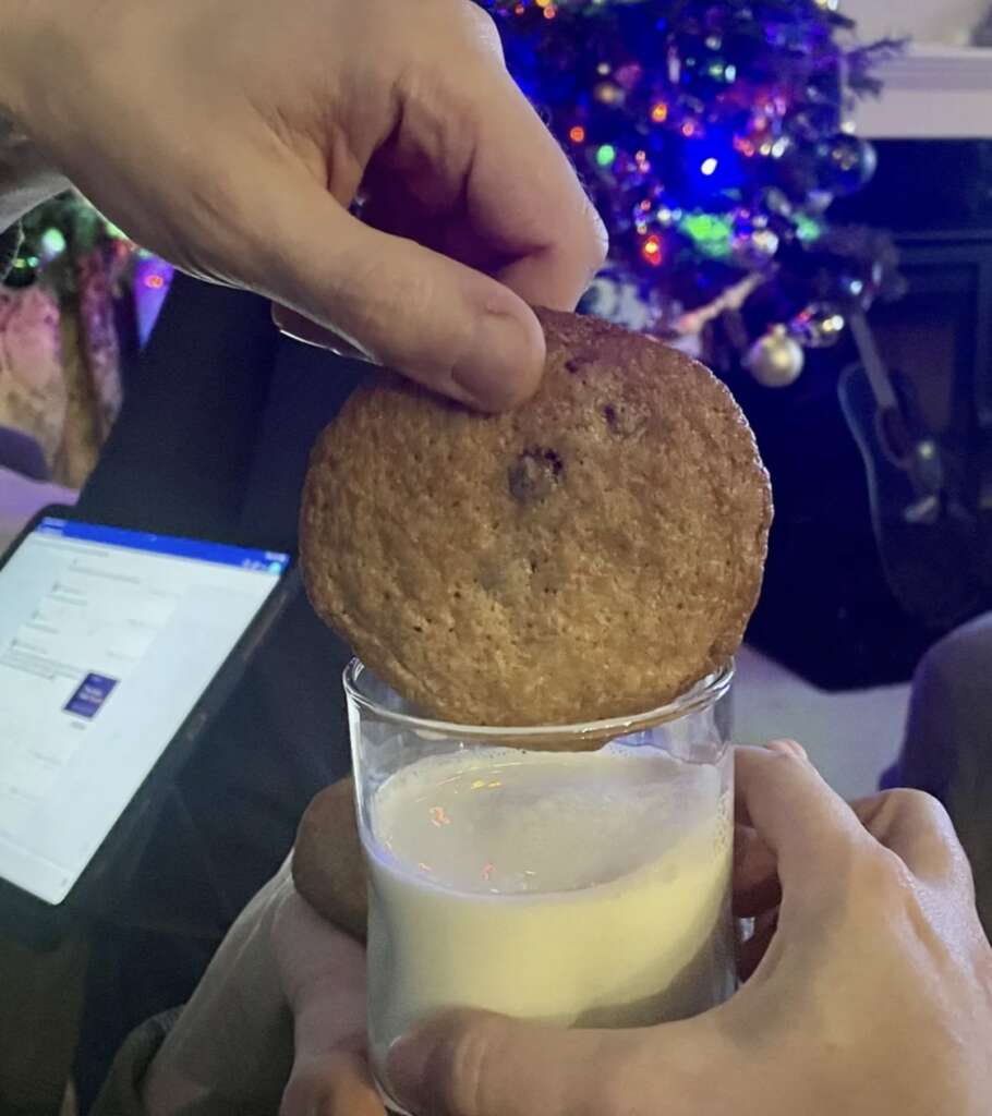 A person dips a chocolate chip cookie into a glass of milk with a mildly infuriating lack of urgency, as the background features a decorated Christmas tree and lit fireplace. Another person holds the glass steady, while a tablet with an open document sits forgotten on the side.