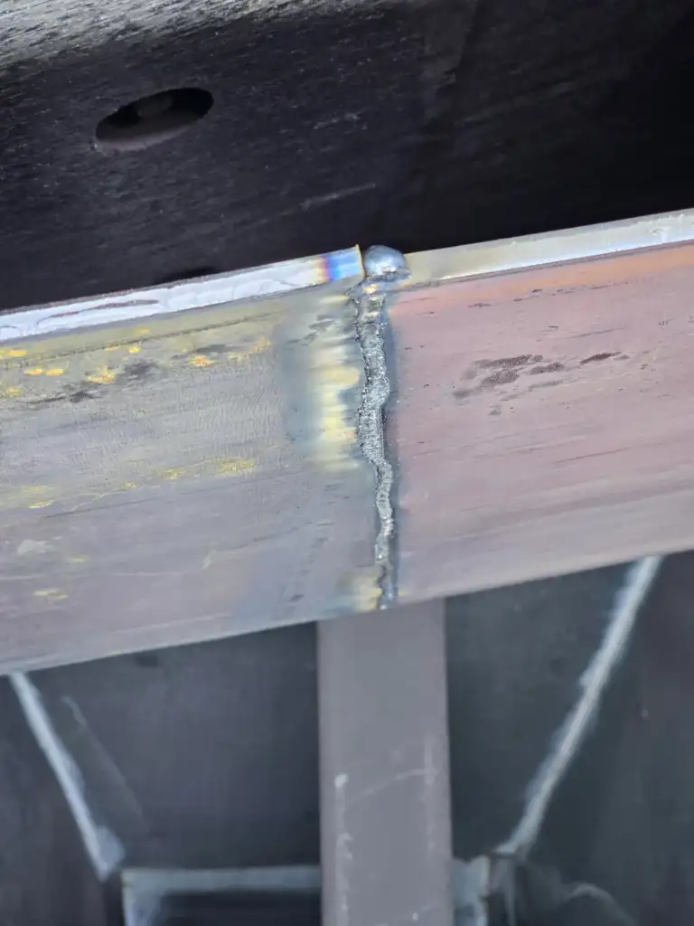 Close-up image of a metal structure showing a welded seam. The weld is slightly uneven with discoloration, transitioning from blue to yellow and silver hues. The background reveals parts of the metal frame with an industrial appearance.