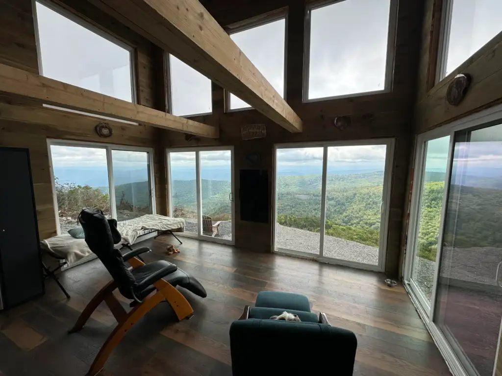 A cozy room with wooden beams and large windows offering a panoramic view of a lush green mountain landscape. It features a reclining chair, a footrest, a small table, and a pet bed, creating a serene and inviting atmosphere.