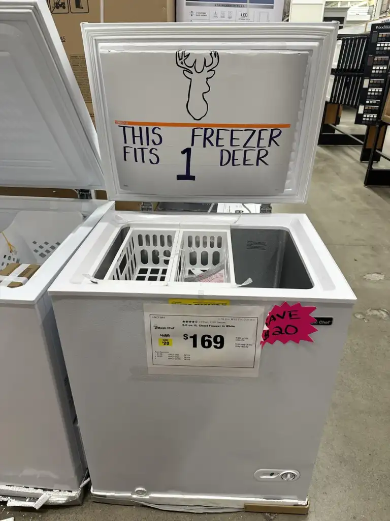 Open white chest freezer with a sign on the lid reading "This Fits 1 Deer" and a deer head drawing. Price tag below shows $169 with a $30 discount. The freezer is in a store aisle.