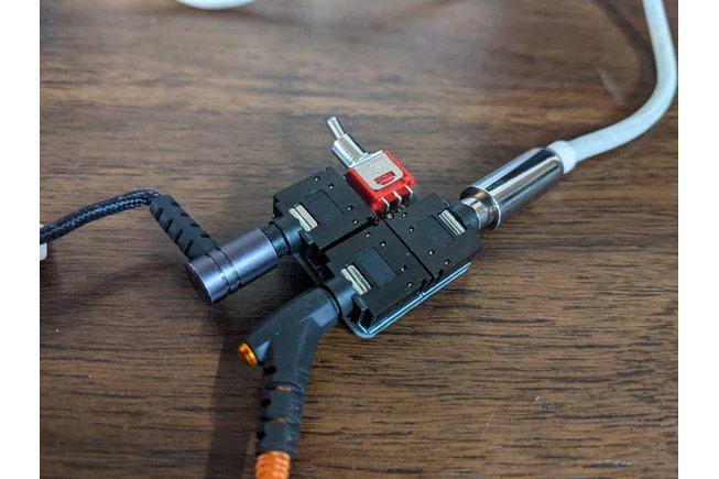 Close-up of an audio setup featuring a red toggle switch connected to two black modular plugs and a silver audio connector. The device rests on a wooden surface with various cables extending outwards.