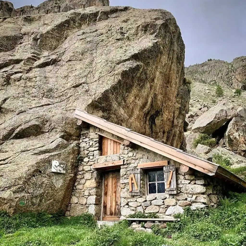 A small stone cabin is nestled against a large rock formation in a rugged landscape. It has a wooden door, a window with wooden shutters, and a grassy foreground. The sky above is overcast.