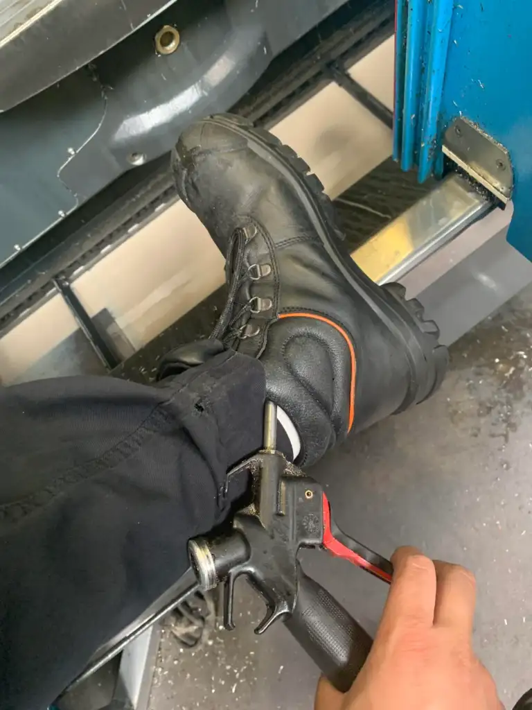 A person wearing black safety boots is using a pneumatic tool to clean or adjust something on a metal step. The tool is held in their hand, and the scene is set inside an industrial or mechanical environment, with blue equipment visible.