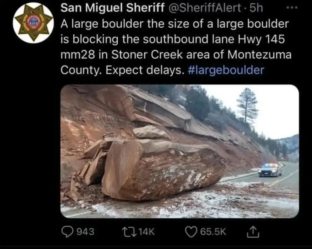 A large boulder blocks the southbound lane of a highway in Montezuma County. A police car is stopped near the boulder, and the road is covered with debris from the landslide. Pine trees and a cloudy sky are in the background.