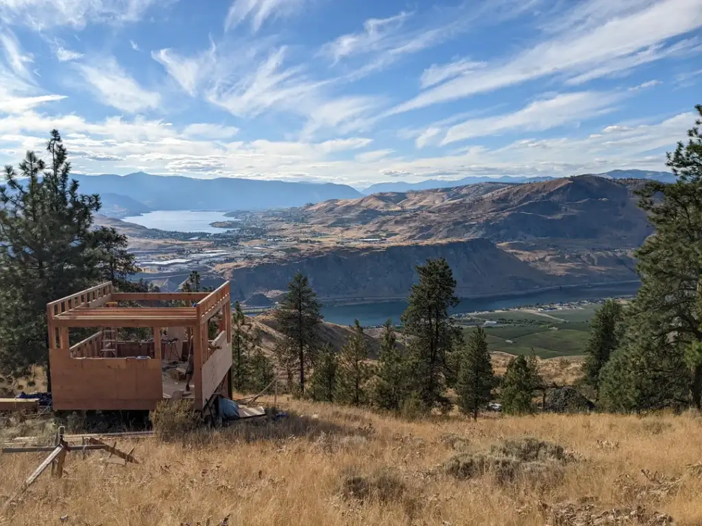 A wooden frame of a cabin under construction sits on a grassy hill, overlooking a scenic landscape with a river, a lake, rolling hills, and mountains under a partly cloudy blue sky. Pine trees surround the area.