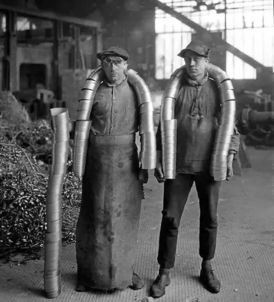 Two workers in a factory, each wearing long metal tubes over their shoulders, resembling armor. They stand in a workshop with tools and machinery in the background. The image is black and white, suggesting it's from an earlier time period.