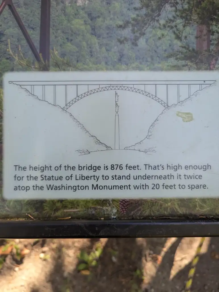 A diagram illustrates a bridge spanning 876 feet high. It shows that the height is enough for the Statue of Liberty to fit underneath it twice, along with 20 extra feet atop the Washington Monument. Text and trees are visible in the background.