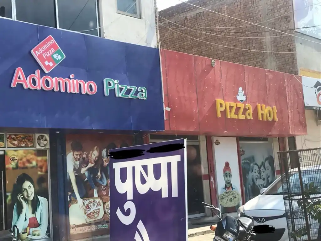 A street view of two adjacent storefronts: "Adomino Pizza" with a blue sign and "Pizza Hot" with a red sign. Both shops display food images. A motorcycle is parked in front, and a purple sign is partially visible.