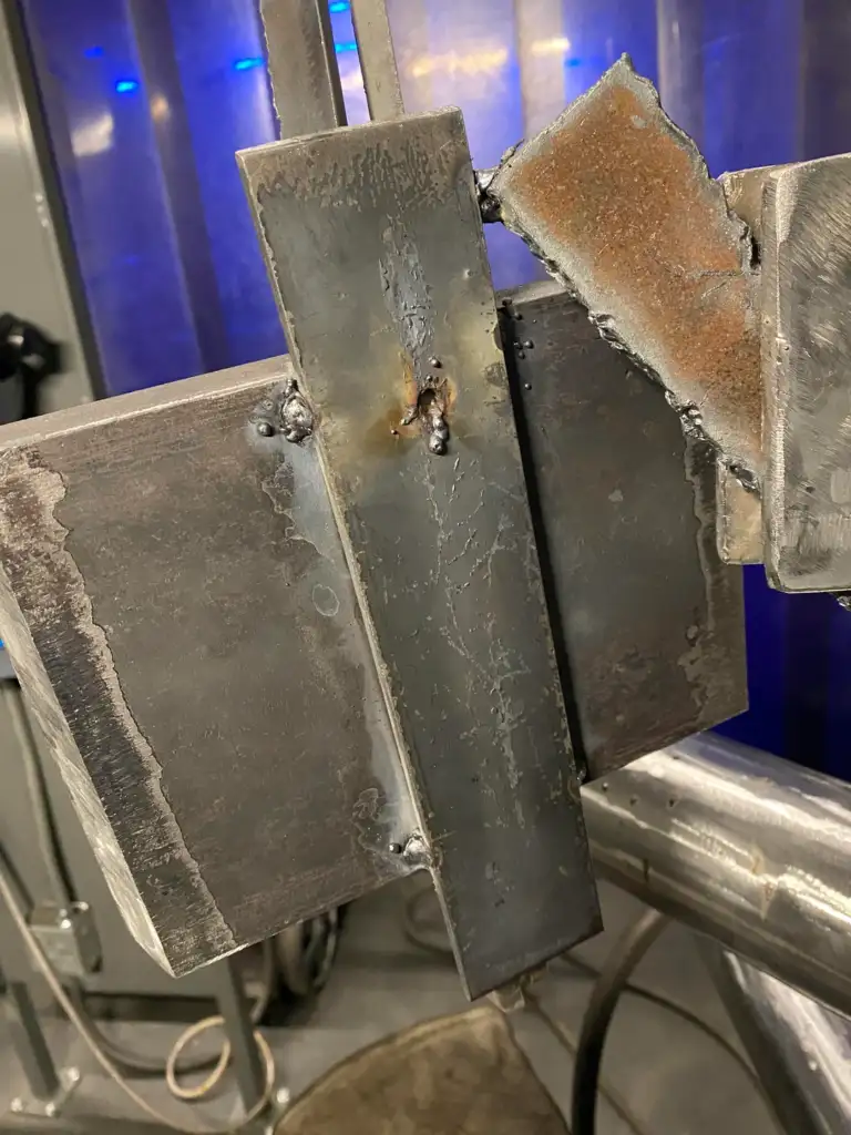 A close-up of a metal welding project showing three intersecting metal beams. The welds are visible, with some discoloration around the joints. The background includes industrial equipment and a blue-tinted curtain.