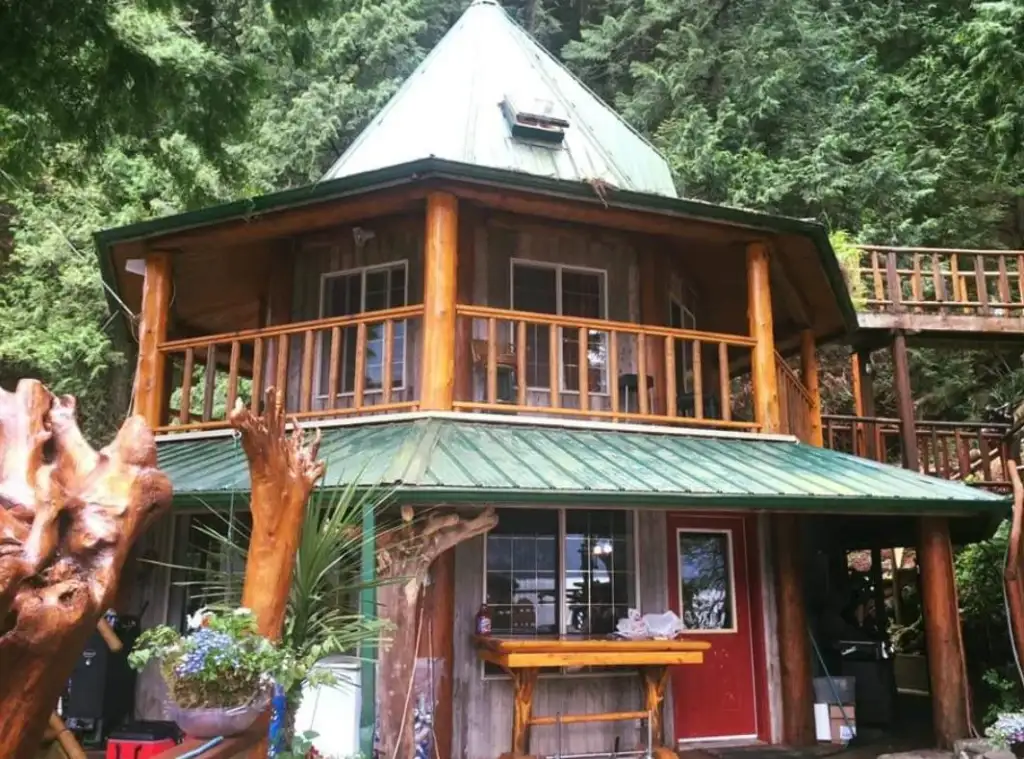 A two-story wooden cabin with a green roof and wraparound balcony is nestled among lush trees. The ground level features a red door and large windows, and there are wooden sculptures and plants in the foreground.