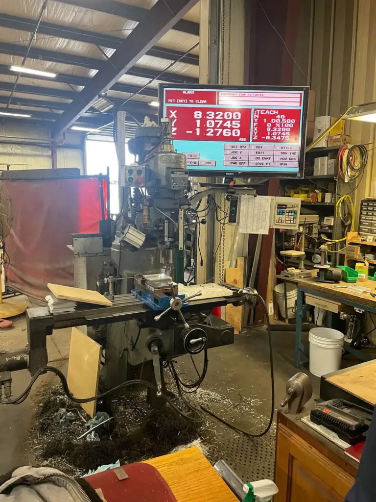 A milling machine in a workshop with metal shavings scattered on the floor. A digital display above shows measurements. Surrounding the machine are workbenches with tools and materials, under a metal roof with beams.