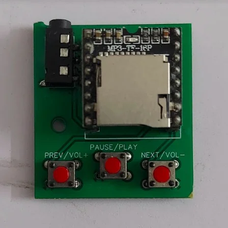 A small electronic circuit board featuring a memory card slot labeled MP3-TF-16P. It includes three red buttons labeled "PREV/VOL+", "PAUSE/PLAY", and "NEXT/VOL-".