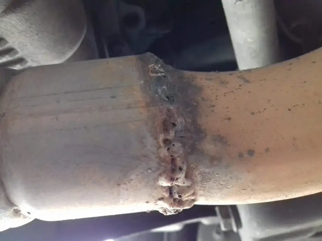 A close-up of a metal pipe with a welded joint. The welding is uneven with visible slag and discoloration on the surface. The pipe appears to be part of a mechanical or automotive system, surrounded by other metal components.