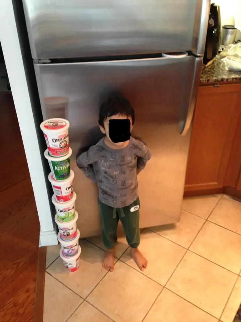 A child stands indoors next to a tall stack of empty yogurt containers reaching up to the refrigerator handle. The child's face is obscured, and they are barefoot on a tile floor with a wooden cabinet to the right.