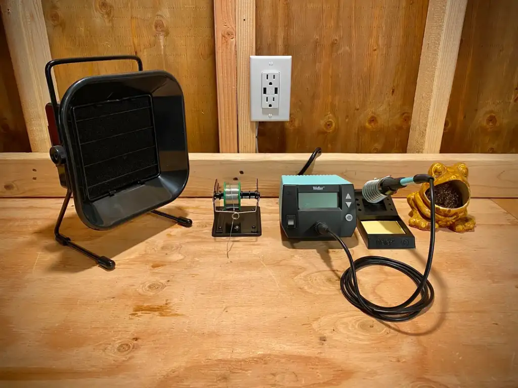 A soldering workstation sits on a wooden surface, featuring a soldering iron with a digital station, a black fume extractor, a circuit board holder with components, and a decorative statue resembling a creature with an open mouth nearby.