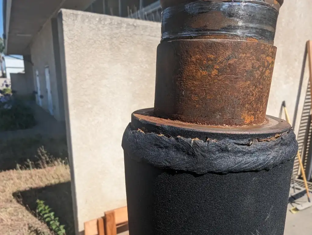 A close-up of a rusted metal pipe wrapped with black insulation material. The pipe shows signs of wear and deterioration. The background is blurred, showing a concrete building and some greenery.