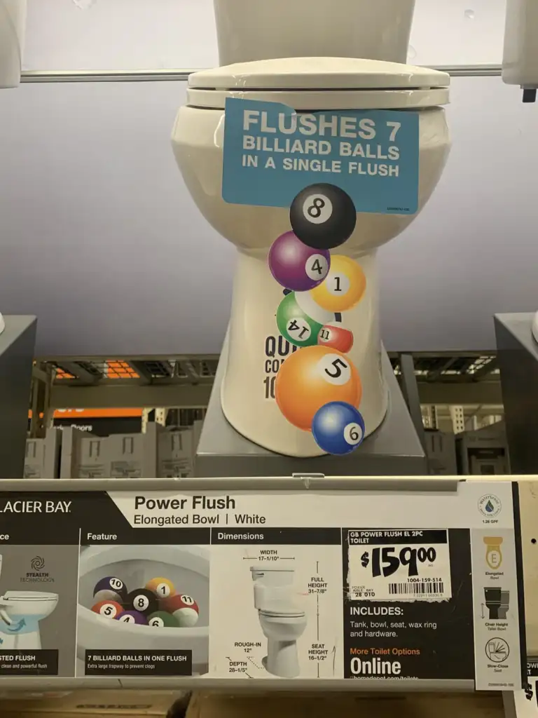 A white toilet is displayed in a store with a sign showing it can flush seven billiard balls in a single flush. The price tag reads $159.00.