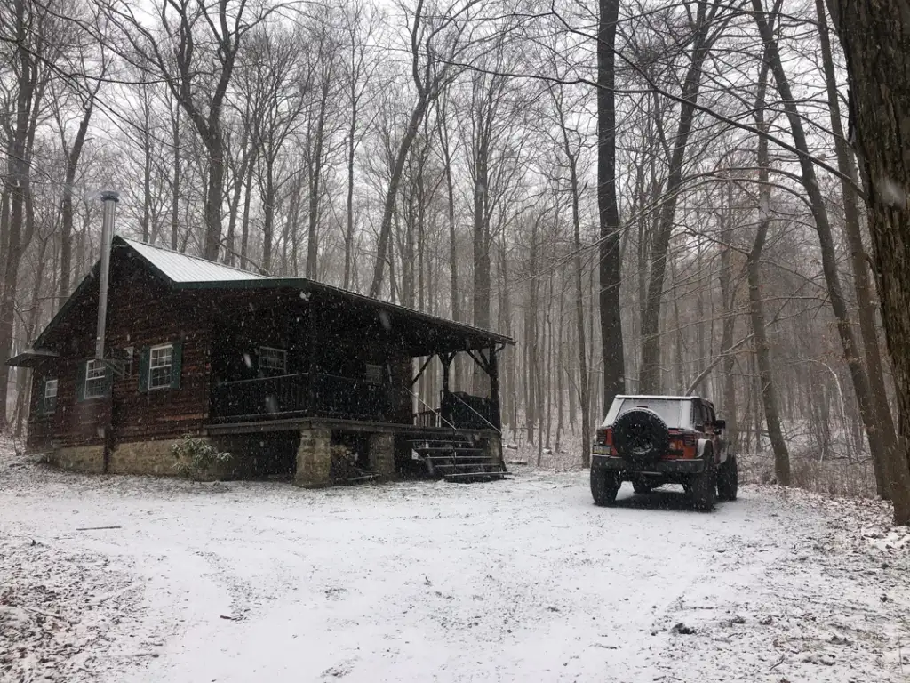 A rustic cabin with a small porch sits in a snowy forest. A vintage red SUV is parked nearby. Bare trees surround the scene, creating a tranquil winter setting. Snowflakes are gently falling, covering the ground in a thin, white layer.