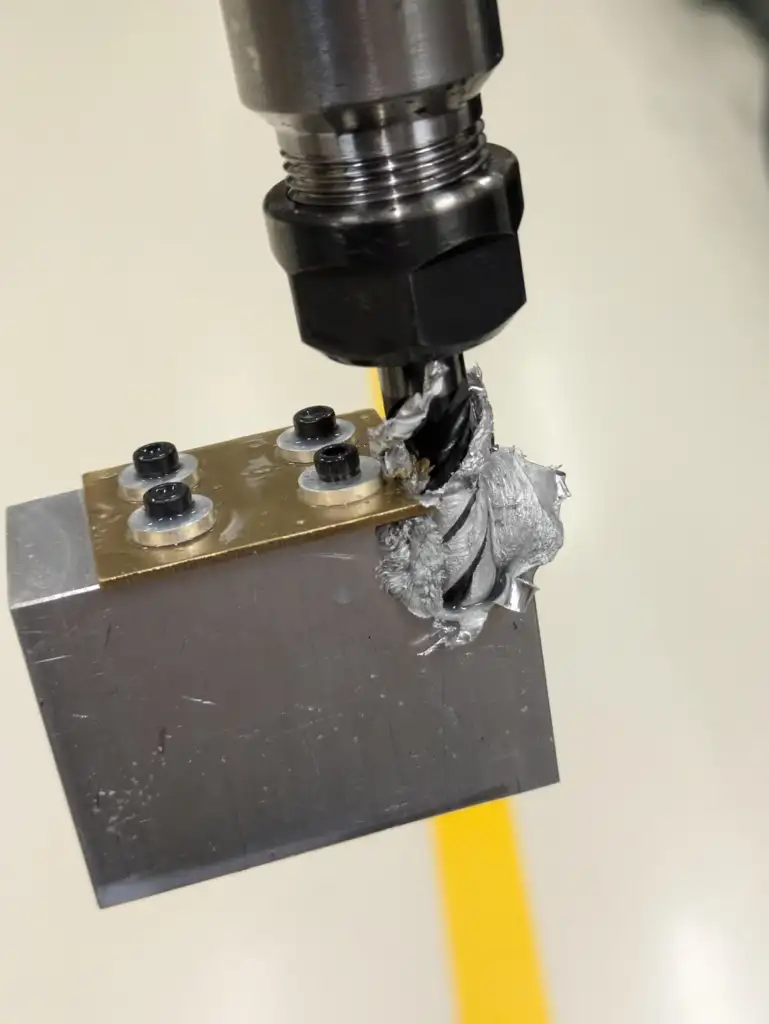 A close-up of a drill bit cutting into a metal block, creating spirals of metal shavings. The block has a metal plate with three bolts on top. The background is blurred, focusing attention on the drilling process.