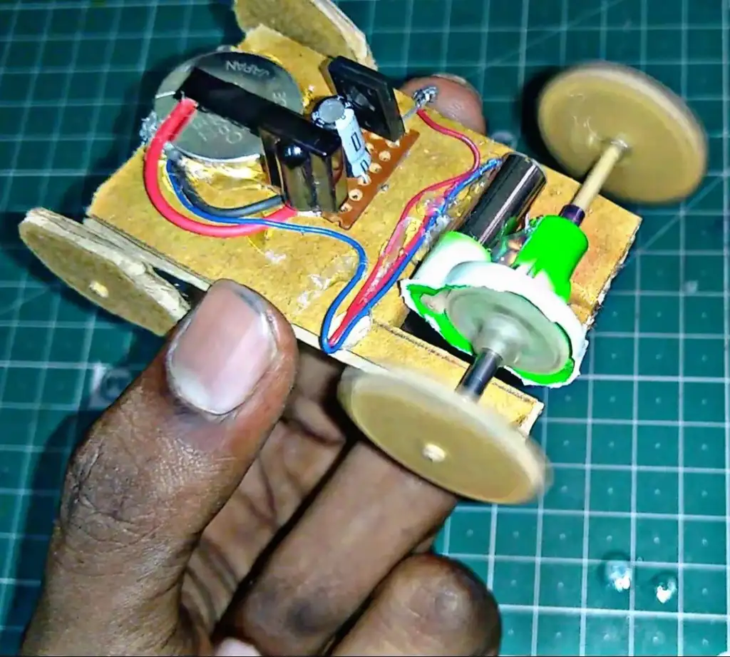 A person holds a small DIY car made from cardboard, with visible wires, a battery, and a small motor. The car is placed on a green cutting mat, and the wheels are made of round discs.