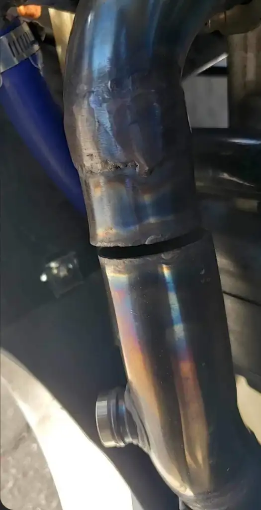 A close-up of a metal pipe with a large crack, showing discoloration from heat. The pipe is part of a larger mechanical assembly, with a blue component visible in the background.