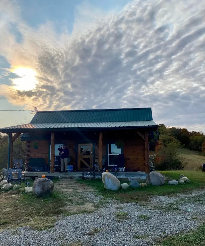 A rustic cabin with a green roof sits under a dramatic cloudy sky. A person stands on the porch, and a bicycle is parked nearby. The yard features large rocks, and trees are visible in the background.