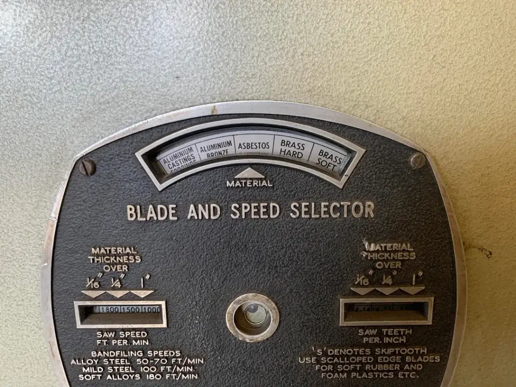 Close-up of a vintage machine's control panel labeled "Blade and Speed Selector." It includes material types like aluminum and brass, and settings for blade speed and thickness. The panel is slightly worn, with metal and textured surfaces.